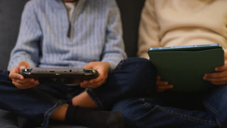 Close-Up-Of-Two-Young-Boys-Sitting-On-Sofa-At-Home-Playing-Games-Or-Streaming-Onto-Digital-Tablet-And-Handheld-Gaming-Device
